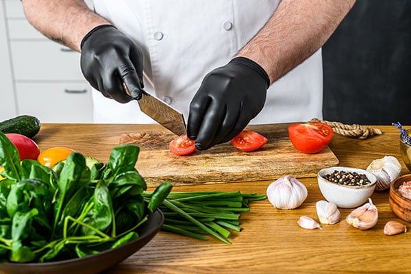 A chef in black gloves is slicing tomatoes on a wooden chopping Board.